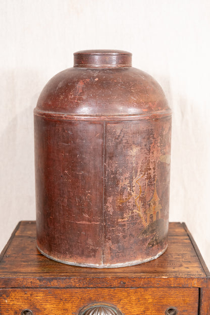 A vintage distressed brown metal canister with a domed lid, displayed on top of a wooden chest against a light off-white background.