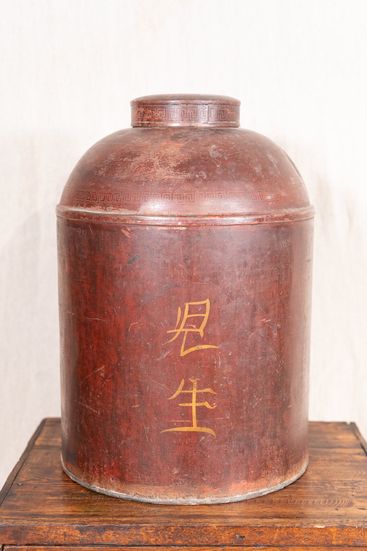 A large cylindrical brown wooden or metal storage jar with gold Chinese characters on the front, displayed on a wooden surface against a light neutral background.