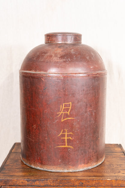 A large cylindrical brown wooden or metal storage jar with gold Chinese characters on the front, displayed on a wooden surface against a light neutral background.