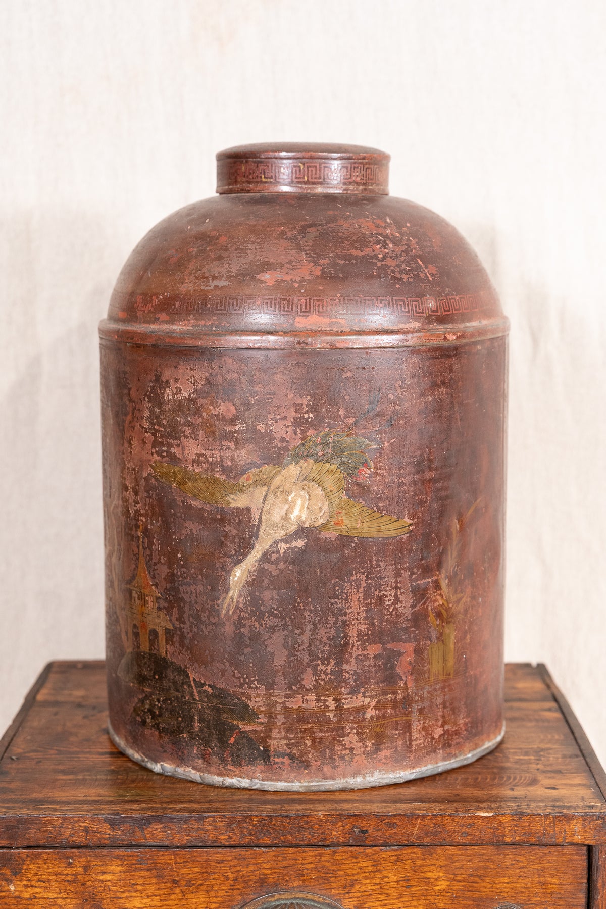 An antique round metal canister with a domed lid, featuring a distressed reddish-brown patina and a faded painted bird design on the front, displayed on a wooden surface against a light textured wall.