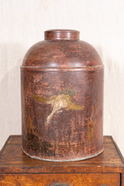 An antique round metal canister with a domed lid, featuring a distressed reddish-brown patina and a faded painted bird design on the front, displayed on a wooden surface against a light textured wall.