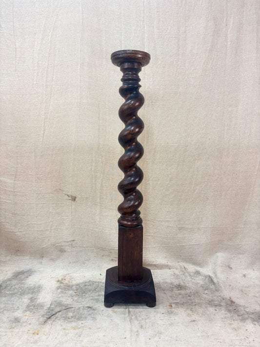 A tall, dark wooden pedestal or plant stand featuring a prominent barley twist central column and a square base.