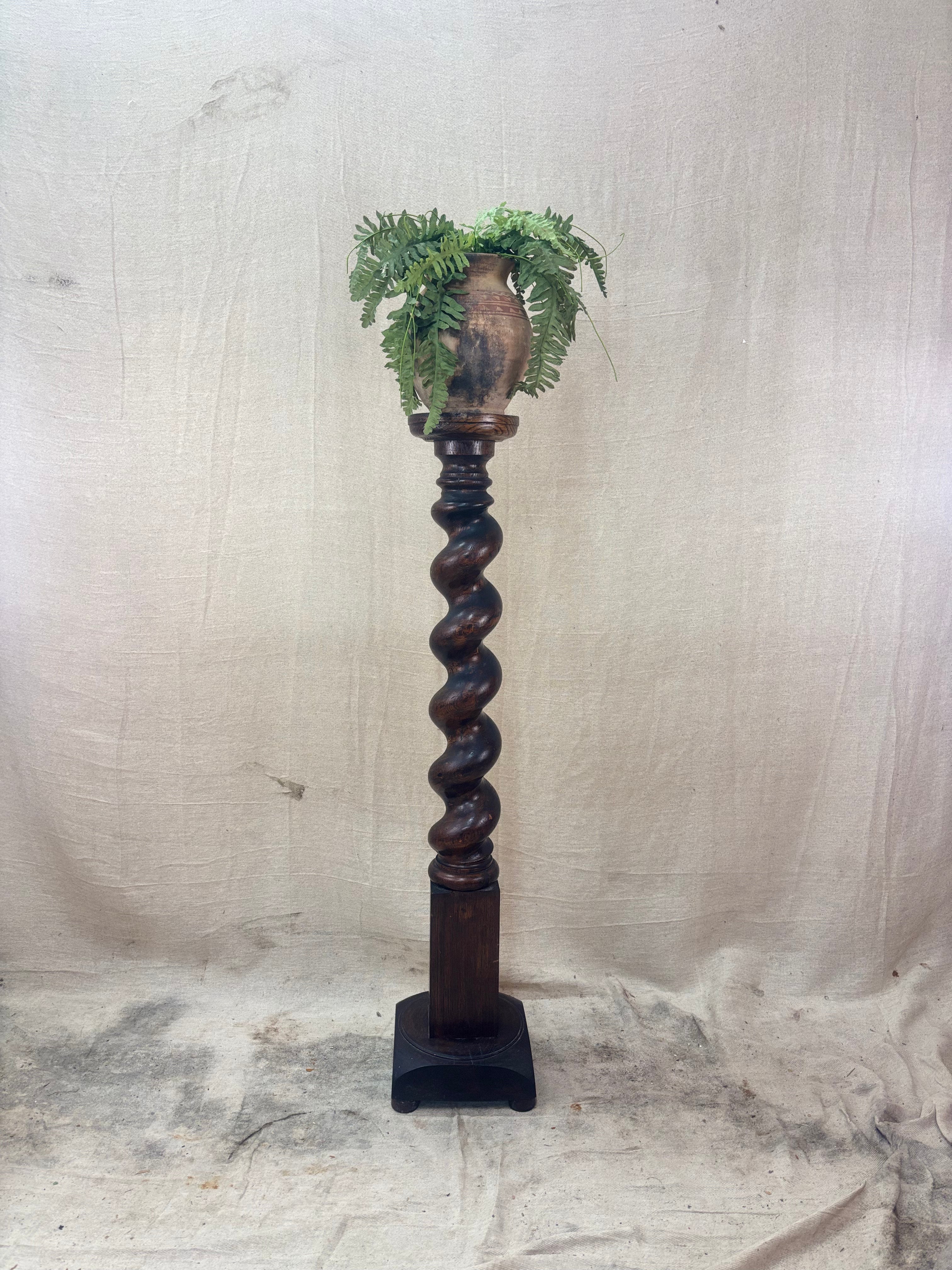 A tall, dark wooden pedestal or plant stand featuring a prominent barley twist central column and a square base.