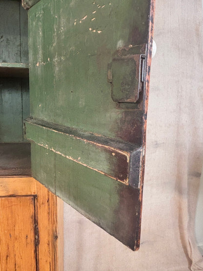 Close-up of a distressed green wooden cabinet door with a metal latch, showing peeling paint and weathered edges, set against a neutral fabric background.