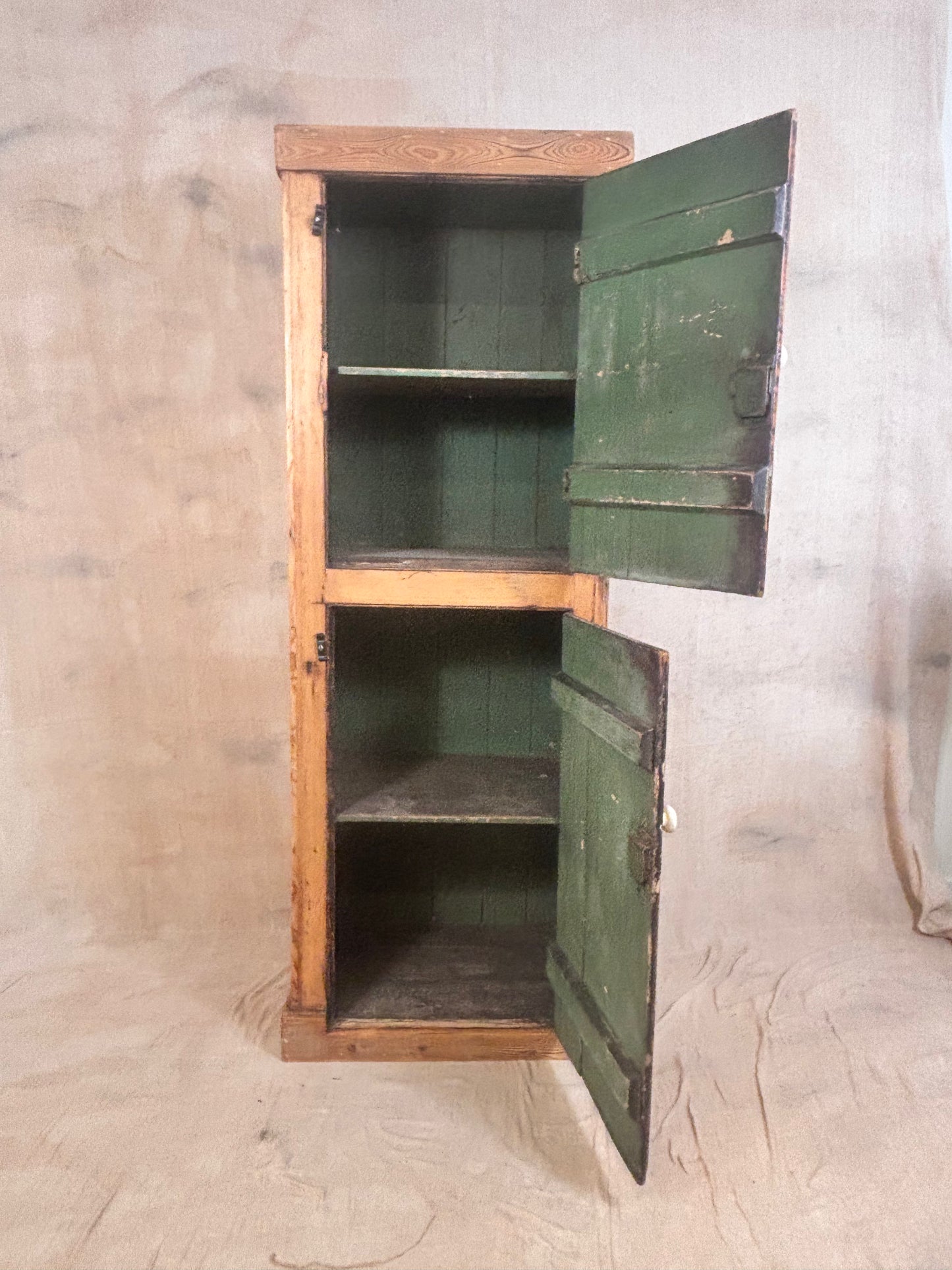 A rustic wooden tall cabinet with two green doors shown open, revealing two interior shelves, set against a neutral fabric backdrop.