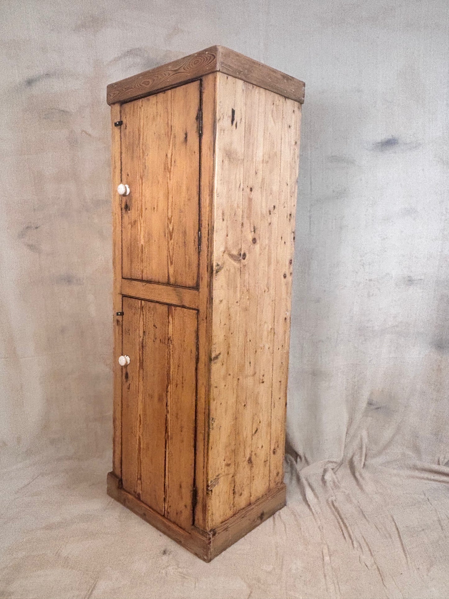 A rustic pine wood storage cabinet with two doors and small white knobs, standing against a draped fabric backdrop.