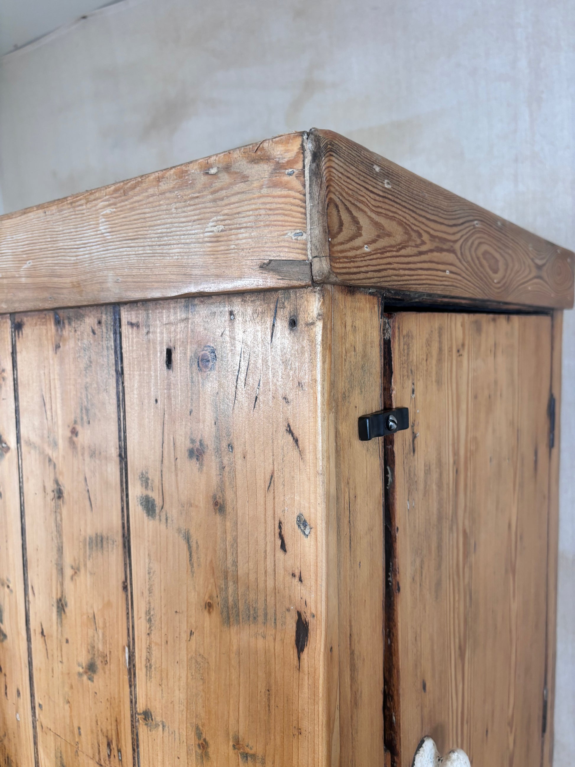 Close-up of a rustic wooden cabinet or wardrobe corner, showing natural pine wood grain, a black metal latch, and a light neutral wall background.