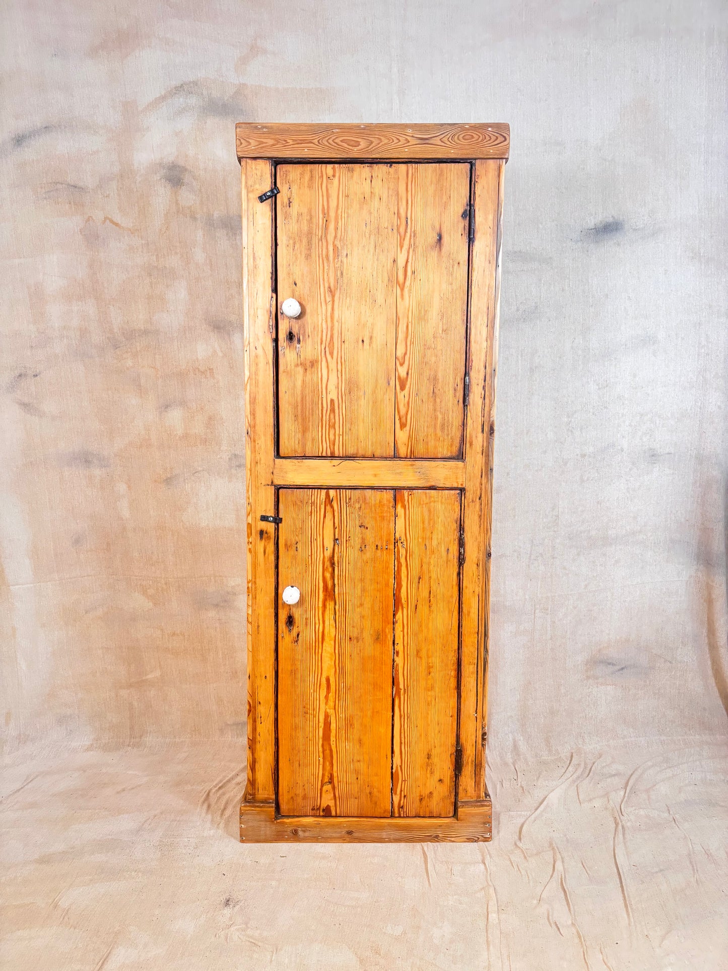A tall, narrow rustic pine cabinet with two doors and small round white knobs, photographed against a beige fabric backdrop.