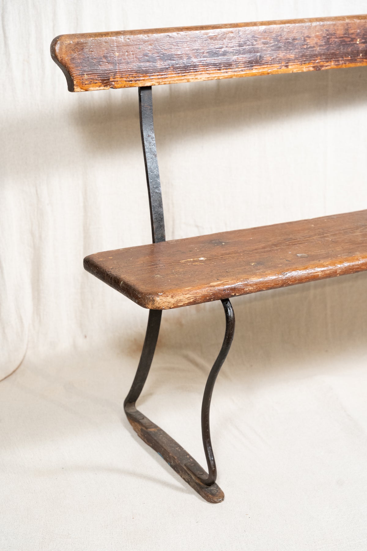 A rustic wooden bench with a weathered brown seat and backrest, supported by curved black metal legs, photographed against a draped off-white fabric backdrop.