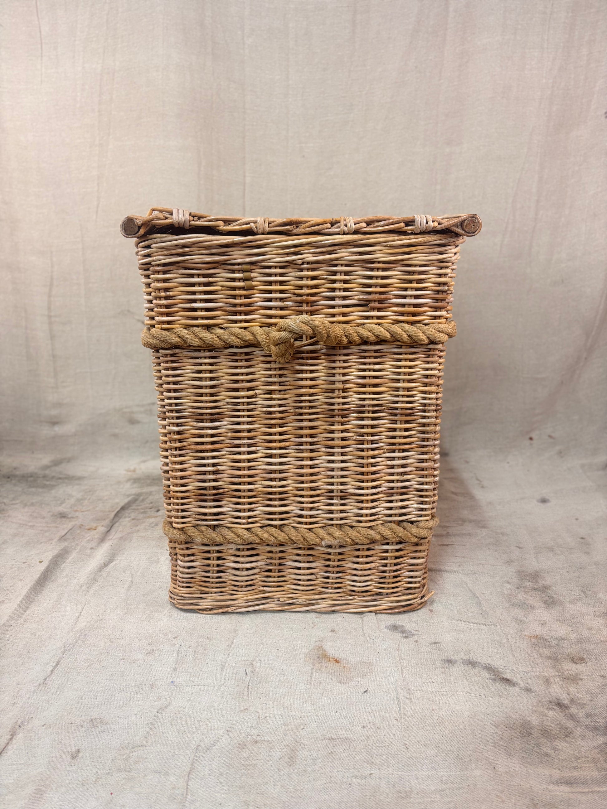 Square wicker storage basket with lid and rope banding, photographed against a neutral backdrop.