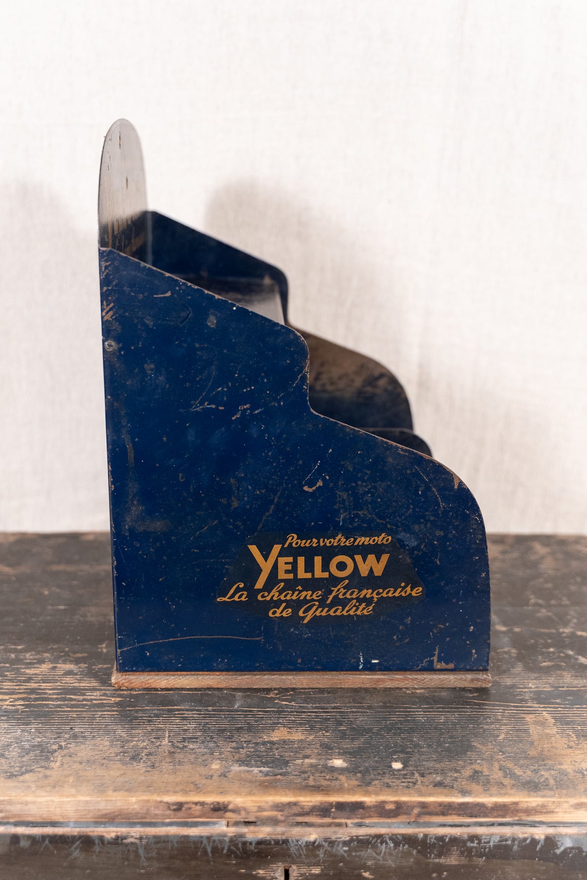 A vintage dark blue metal magazine rack with gold lettering reading 'Yellow' and French text, standing on a rustic wooden surface against a light textured wall.
