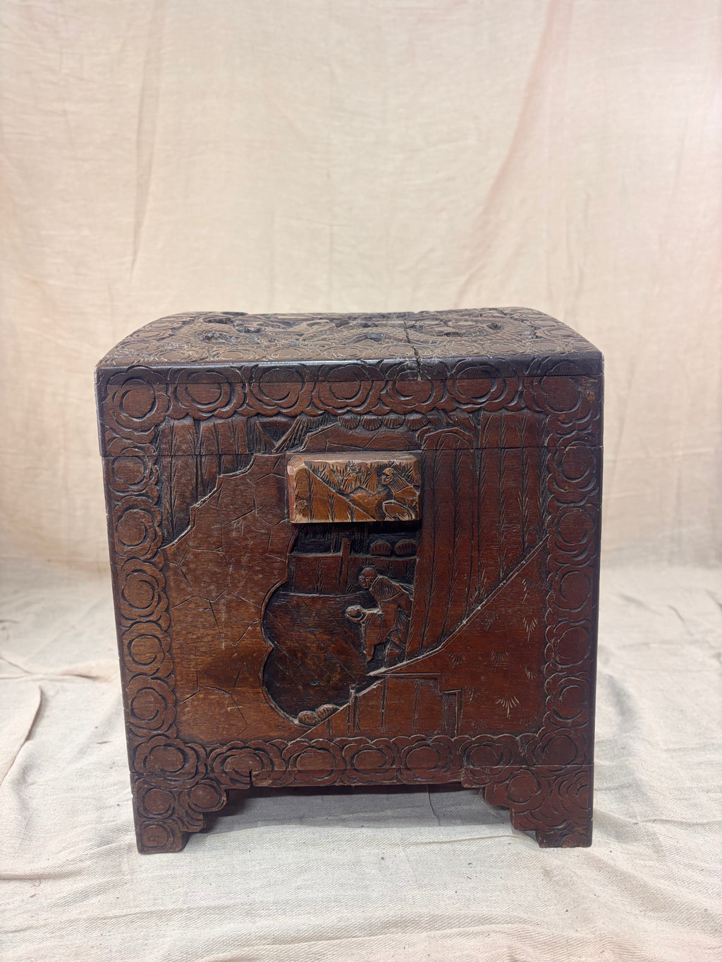 A carved wooden chest with mountain scene and a small wooden handle against a cream cloth