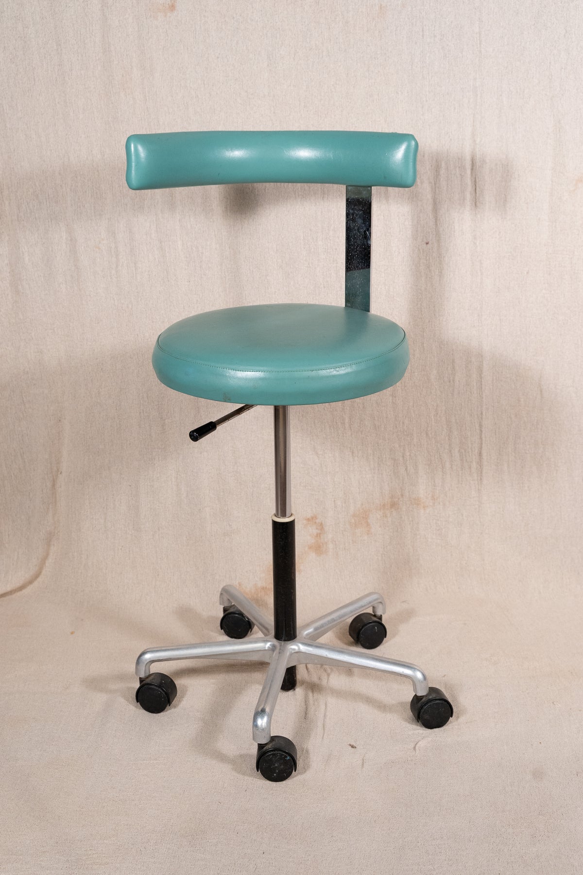 A vintage-style medical or laboratory stool with a rounded teal-green seat and backrest, mounted on a black gas-lift column and a silver five-star wheeled base, photographed against a beige fabric backdrop.