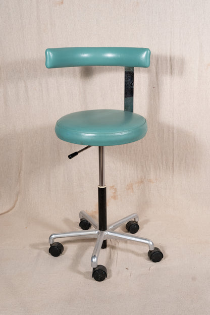 A vintage-style medical or laboratory stool with a rounded teal-green seat and backrest, mounted on a black gas-lift column and a silver five-star wheeled base, photographed against a beige fabric backdrop.