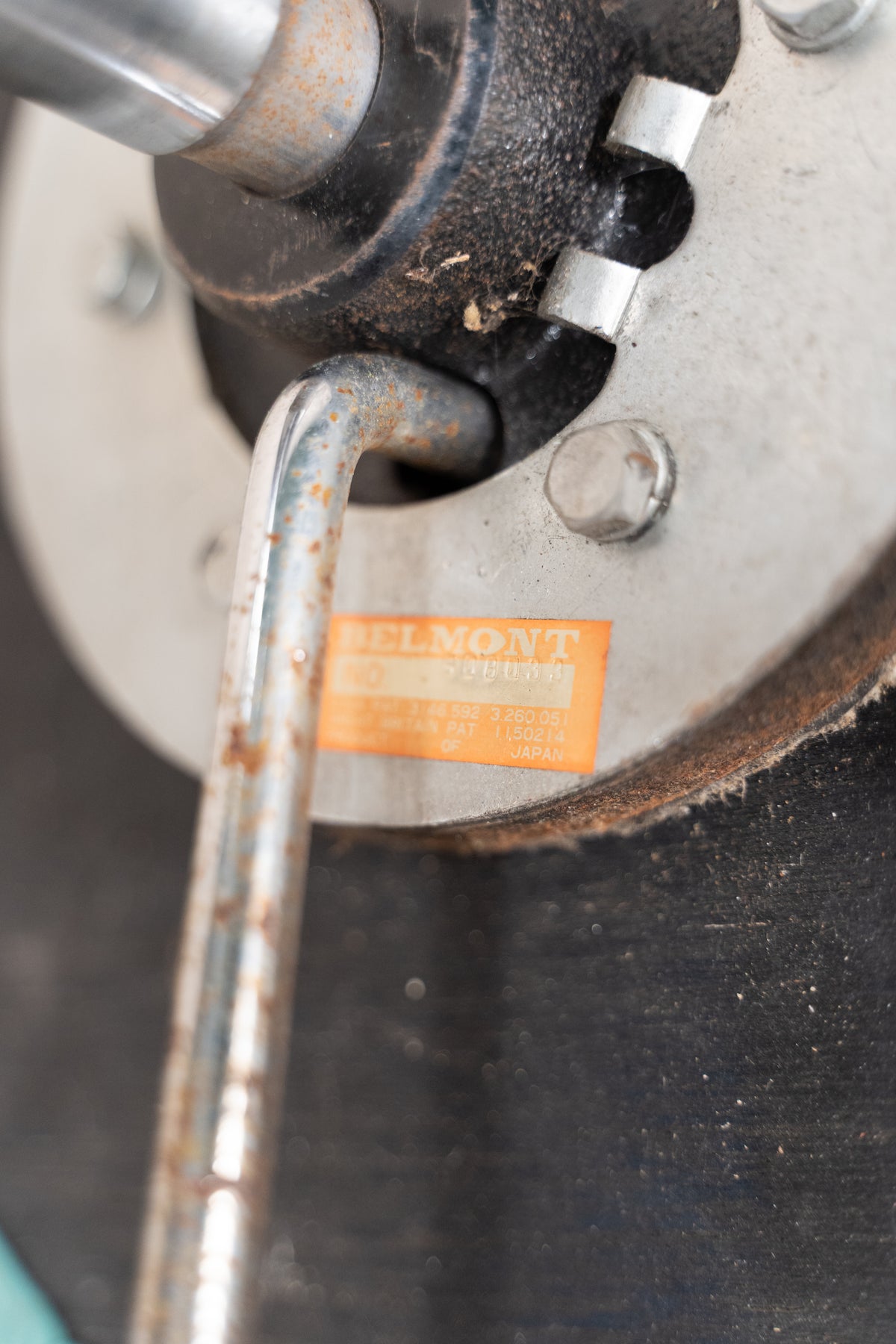 Close-up of a vintage metal machine component with a visible orange label reading 'BELMONT' and a metal tool inserted into a threaded opening.