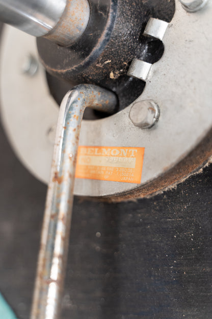 Close-up of a vintage metal machine component with a visible orange label reading 'BELMONT' and a metal tool inserted into a threaded opening.