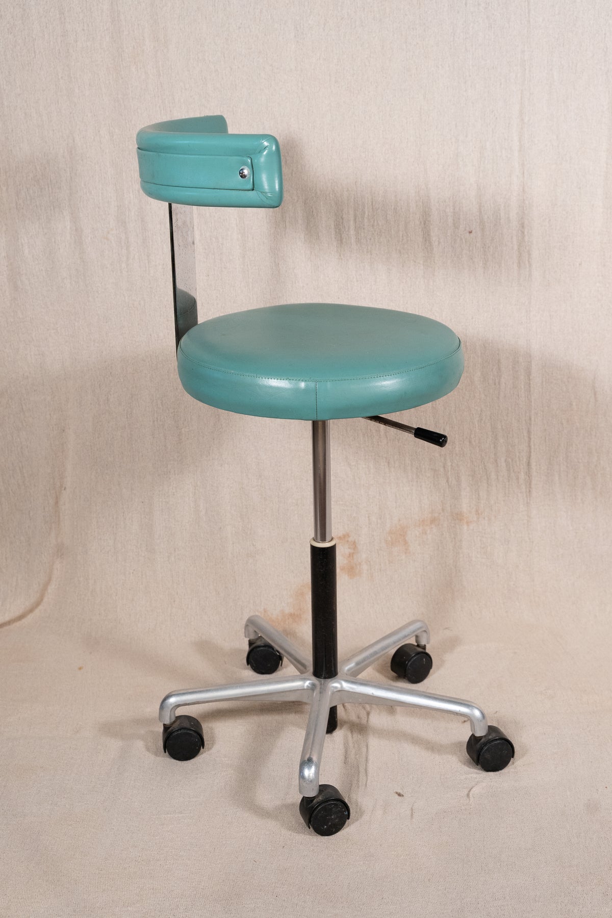 A vintage-style medical or examination stool with a rounded turquoise seat, matching backrest, chrome five-star base, and black caster wheels, photographed against a beige fabric backdrop.