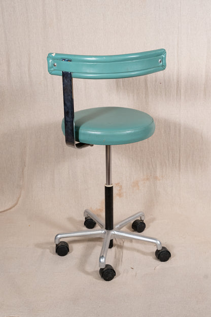 A vintage-style teal office stool with a padded seat, a curved backrest, a black vertical support strap, and a silver five-star wheeled base, photographed against a beige fabric backdrop.