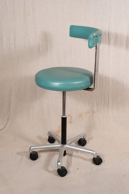 A vintage-style teal medical or laboratory stool with a round upholstered seat, adjustable metal leg, and backrest, shown against a neutral beige fabric backdrop.