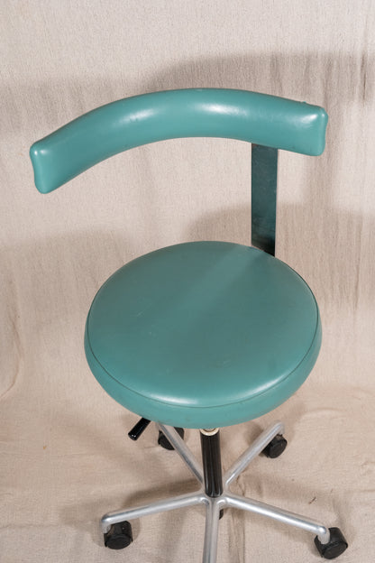 A teal-green swivel stool with a curved backrest, upholstered seat, and a five-star chrome base with black casters, set against a beige fabric backdrop.