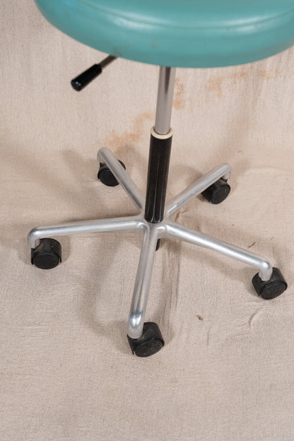A close-up of a vintage-style swivel stool showing its teal seat, chrome five-star base, black casters, and black central column on a beige fabric background.