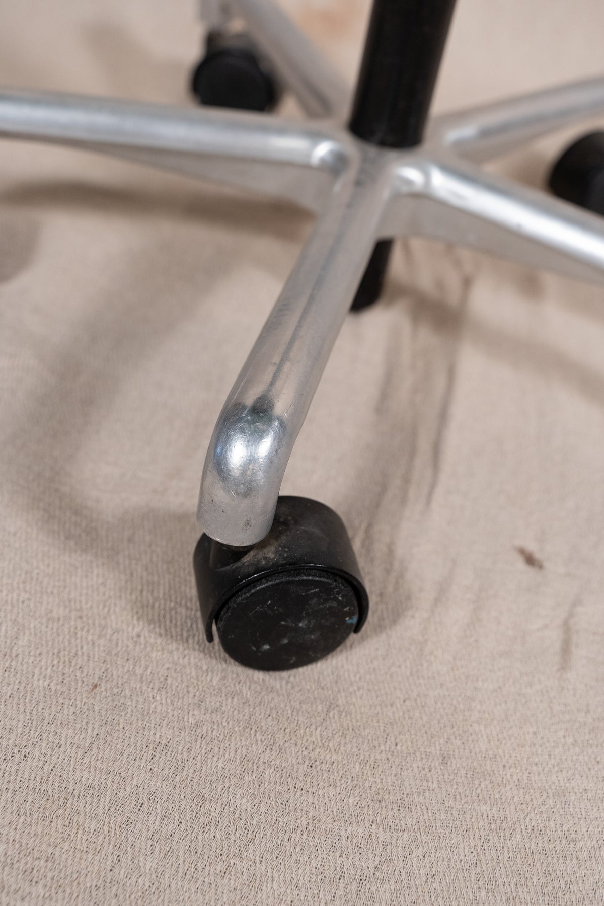 Close-up of a black caster wheel attached to a silver metal five-star chair base, resting on a beige fabric surface.