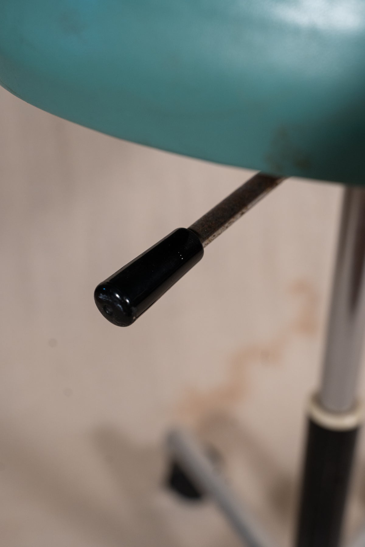 Close-up of a black adjustable stool leg with a metal rod and a teal-colored seat cushion in the background.