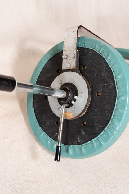 Close-up of a circular teal and black cushioned wheel or pad with a metal central hub and a long metal rod extending from the left, set against a light beige fabric background.