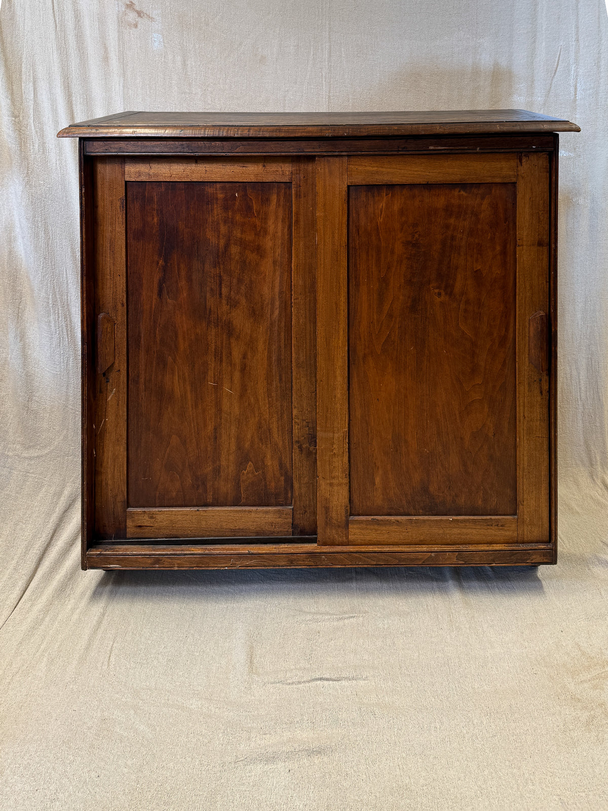 A vintage dark brown wooden cabinet with two paneled doors, photographed against a draped beige fabric backdrop.