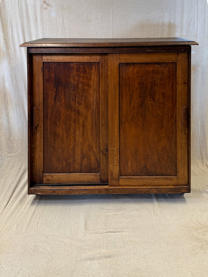 A vintage dark brown wooden cabinet with two paneled doors, photographed against a draped beige fabric backdrop.