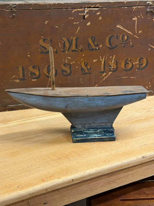 A vintage wooden toy boat with a blue-gray painted finish and a small wooden sail, displayed on a light wood tabletop in front of a rustic brown wooden box with 'S.M.