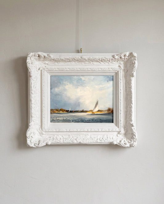 A framed landscape painting of sailboats on water, displayed in an ornate white frame hanging on a light gray wall.