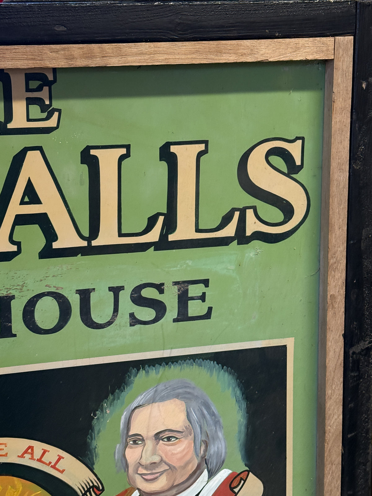 A close-up of a framed vintage-style sign with a light green background, large cream lettering, and a painted portrait at the bottom.
