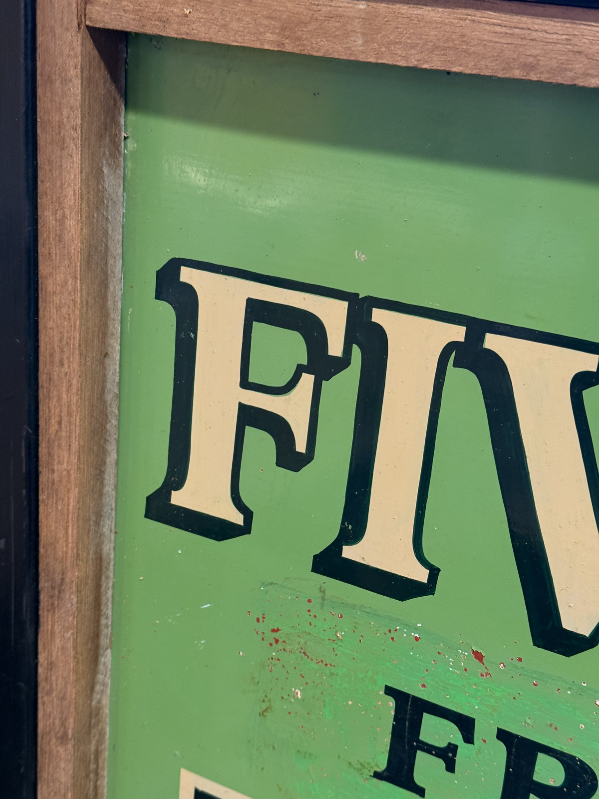 A close-up of a vintage-style green sign with large cream lettering outlined in black, partially showing the word 'FIVE' and other letters, bordered by a wooden frame.