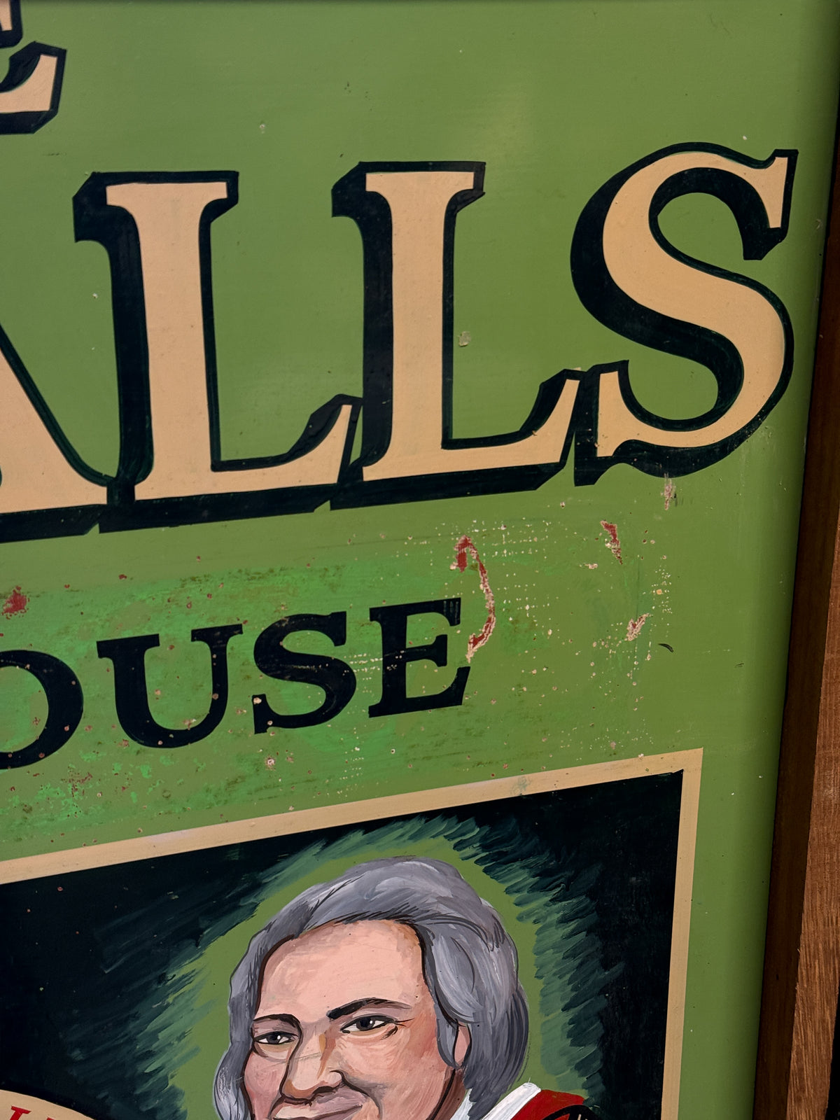 Close-up of a vintage-style painted sign with a green background, large tan lettering outlined in black, and part of a painted portrait at the bottom.